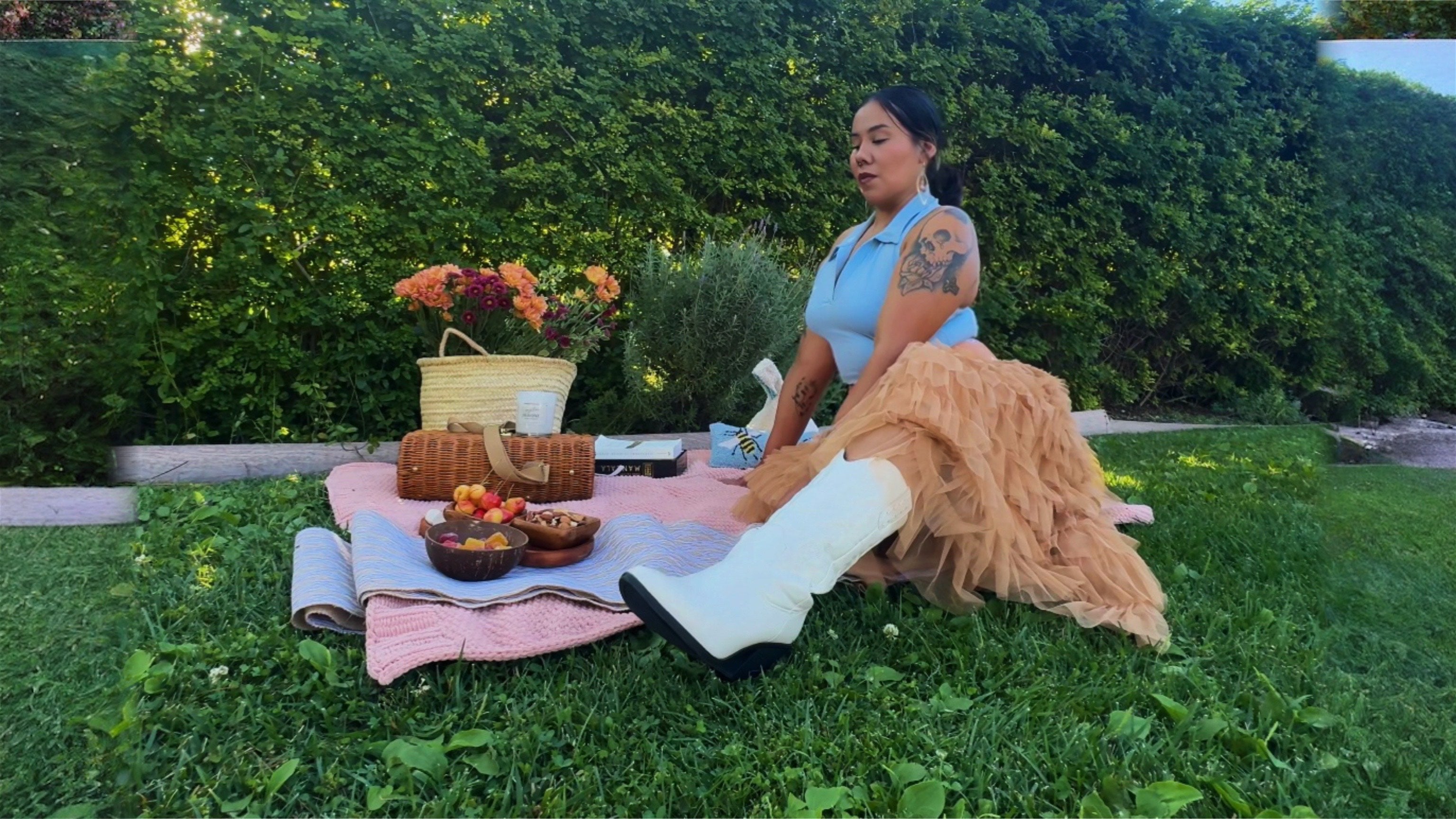 Load video: Indigenous female, baddie sitting in a park having a picnic for herself. Living her best life. Sitting with fresh flowers, fruit, nuts and cherries. Wearing a light blue short sleeve polo, frilly long skirt, cowgirl boots and reading Braiding Sweetgrass by Robin Wall Kimmerer.